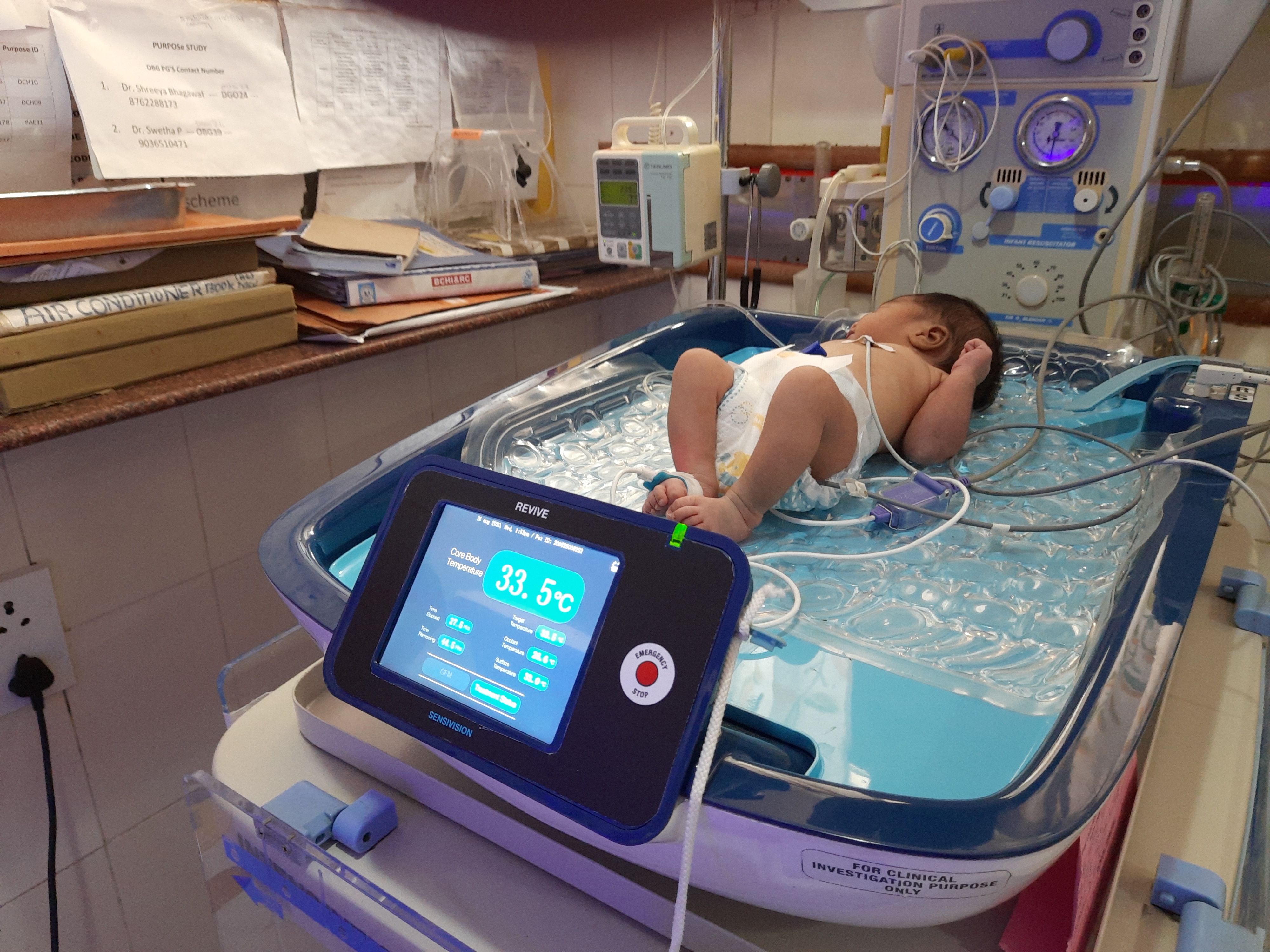 Clinical REVIVE implementation showing newborn baby on cooling system with clear blue cooling mattress, prominent touchscreen display showing 33.5°C temperature reading, multiple monitoring parameters, REVIVE branding with Sensivision logo, hospital NICU environment with medical equipment and supplies visible in background